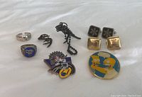 Photo showing all items laid out on white fabric: 2 rings, 4 cufflinks, 3 lapel pins, and dinosaur charm keychain.