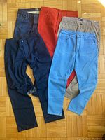 Five pairs of men's pants displayed flat on parquet floor, variety of colors including dark navy, bright red, beige, and shades of blue, showing general shape and style of pants.