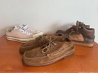 Three pairs of men's shoes: beige Converse low top sneakers, brown Sperry suede boat shoes, and two-tone brown Alife hi-top sneakers.