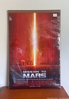 Full view of the Mission to Mars movie poster with plastic covering, showing red and orange hues and central light beam.