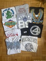 Top-down view of ten assorted vintage men's T-shirts laid flat, showcasing various graphics and characters including MGM, Toronto Maple Leafs, Gemini zodiac sign, eagle illustration, and cinema promo shirts.