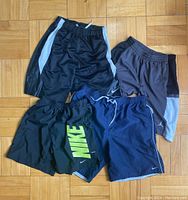 Four pairs of Nike shorts arranged on wooden floor showing different styles and colors including black with white sides, blue with piping, and black with large green Nike text and Jordan shorts.