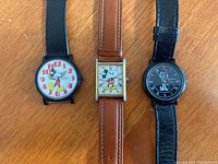 Three wristwatches on a wooden surface: two Disney-themed (Mickey Mouse) and one Felix the Cat watch