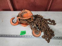 Full view of the orange chain operated hoist with attached heavy rusted chain and large hooks resting on a flat surface with a ruler beside measuring length.