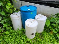 Five dock float barrels positioned on grass by a dock. One large blue barrel stands in the back with four smaller white barrels in front. The barrels have dirt and wear consistent with outdoor use.