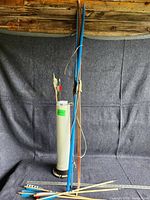 Full view of three bows standing upright alongside white quiver with arrows inside, set against a cloth backdrop and wood wall.