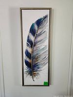Full front view of framed feather artwork hung on white wall, showing entire feather and frame.