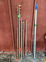 Five metal bar clamps leaning against a red-painted wooden wall showing varying lengths and visible rust and wear.