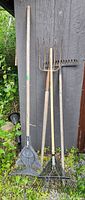 Four garden hand tools leaning against a wooden wall outdoors, including a plastic leaf rake, two metal rakes, and a 5-tine metal pitchfork with wooden handles showing rust and wear.