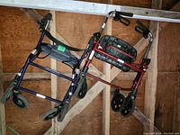 Two folding walkers leaning against a wooden wall. One blue, one red with black fabric basket.