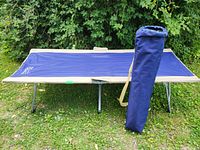 Blue Easy Cot camping cot with beige edges and blue carry bag standing upright next to it on grass.