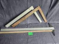 Set of measuring tools including T-square, roofing squares, and yardstick laid on dark cloth