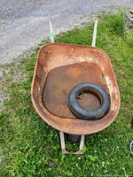 Rusty metal wheelbarrow with flat tire inside the tub, containing a replacement rubber inner tube, resting outdoors on grass and gravel.