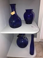 Four blue glass pieces displayed on a white shelf and floor showing different shapes and sizes.