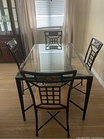 Four metal dining chairs arranged around glass dining table showing black frames with geometric backrest design and beige upholstered cushions.