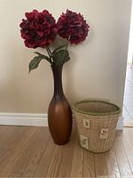 Photo showing entire vase with faux flowers next to woven trash bin on wooden floor against wall