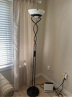 Full view of the black metal torchiere floor lamp with twisted stand and frosted glass shade at top, placed next to beige curtain and wall.