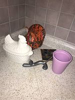 Photo showing a white lidded chicken-shaped ceramic container, an orange-brown turkey ceramic container, a small colorful square plate, a grey metal faucet handle, and a lavender flower pot corner.