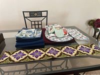 Photo showing folded blue and floral patterned linens, oven mitts, pot holders, and a long fabric wall hanging spread on table.
