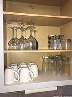Shelf with 6 inverted wine glasses, 3 small glass jars or tumblers with metallic lids, and 5 clear drinking glasses.
