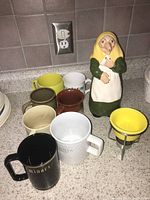 Photo showing seven assorted mugs, ceramic figurine, and yellow butter melter on kitchen counter.