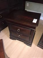 Front and top view of a wooden nightstand showing two drawers with metal ring pulls and dark wood finish.