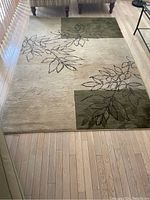 Full view of rectangular area rug on wood floor, showing beige base with green blocks and dark leaf outlines