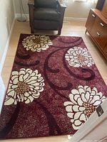 Top angled view of the rectangular burgundy area rug with large beige and brown floral patterns and curved swirls, laid on light hardwood floor near furniture.