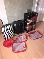 Photo showing three suitcases standing upright: a zebra-striped hard shell, black fabric, and floral hard shell along with four red and gray mesh zip-up packing bags laid flat on floor.
