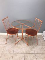 Front view of round metal table with glass top and two matching metal chairs with upholstered brown cushions on tiled floor against gray wall
