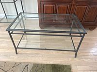 Metal and glass rectangular coffee table with clear surfaces and dark metal frame on wooden floor beside a rug and cabinet.