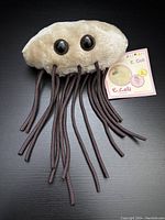 Front view of the beige E. Coli plush toy with multiple brown string appendages and attached tag showing product image and name