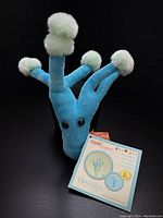 Front view of blue plush toy with white pom-poms on arms and tags showing product info and branding.