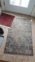 Photo showing small dark red rug with fringe and larger faded beige and gray rug side by side on tiled floor.