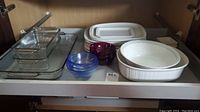 View of several Pyrex and Corning glass baking dishes in clear glass, oval white ceramic baking dish, small colored glass bowls and white lids or baking dishes inside a wooden cabinet drawer.