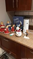 Photo of four ceramic angel candle holders in red and blue robes holding candles, two glass ornaments, a white tea light cover, and a Goebel skier figurine on a kitchen counter with 'Starry Night' box in background
