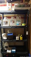 Full shelving unit holding the lot's items including jugs of cleaning fluid, plastic bags, gloves, laundry detergent, and furnace filters on top shelf.