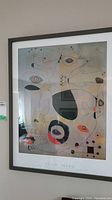 Framed print displayed on the wall showing Joan Miró artwork 'The Port' with abstract shapes and colors, in a glass-covered frame with white mat and a thin wooden frame.