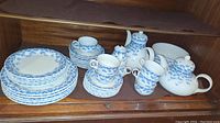 Full view of tea set and plates showing blue floral pattern on white porcelain. Includes stacked plates, cups, cream and sugar pots, and large teapot without lid.