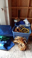 Storage tote with yellow outdoor extension cords and a plumbing U-bend pipe inside, adjacent tote with screws, blue and green tarps stacked on the floor.