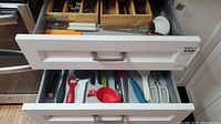 Open drawer showing organized metal cutlery (spoons, forks, knives) in wooden dividers and some larger kitchen utensils in front.