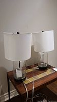 Pair of two table lamps with clear bubble glass cylindrical bases, white fabric shades, on a wooden surface