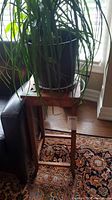 Wide view of vintage wooden plant stand with a dark-colored pot holding a ponytail palm with long leaves hanging down. Shows scratches on wood and surroundings including a patterned rug.