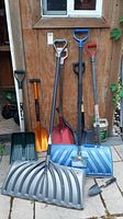 Shows the various shovels and garden tools upright against a wooden shed wall, including three large snow shovels in gray, blue, red, and other smaller shovels.