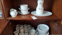 Wide view showing cups, plates, saucers, creamer, sugar bowl, carafe, and bowls on wooden shelves.