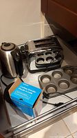 Photo showing all items: stainless steel Hamilton Beach 2-slice toaster, Black & Decker electric kettle, boxed Simplicity coffee grinder, metal muffin tin, and small baking sheet on kitchen countertop.