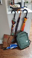 Four umbrellas with straight handles and one compact umbrella alongside a brown floral tote bag and a green Tothemarket backpack sitting on the floor.