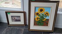 Two framed artworks leaning on floor near a window: a sunflower painting in a large decorative gold frame and a smaller fox print in a simple wooden frame.