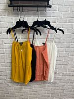 Four tank blouses on hangers against a grey brick wall. Colors visible: mustard yellow, black, dusty rose, white.