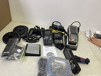 Multiple POS terminal devices including charging docks, cables, handheld scanner, and card readers displayed on a tabletop.